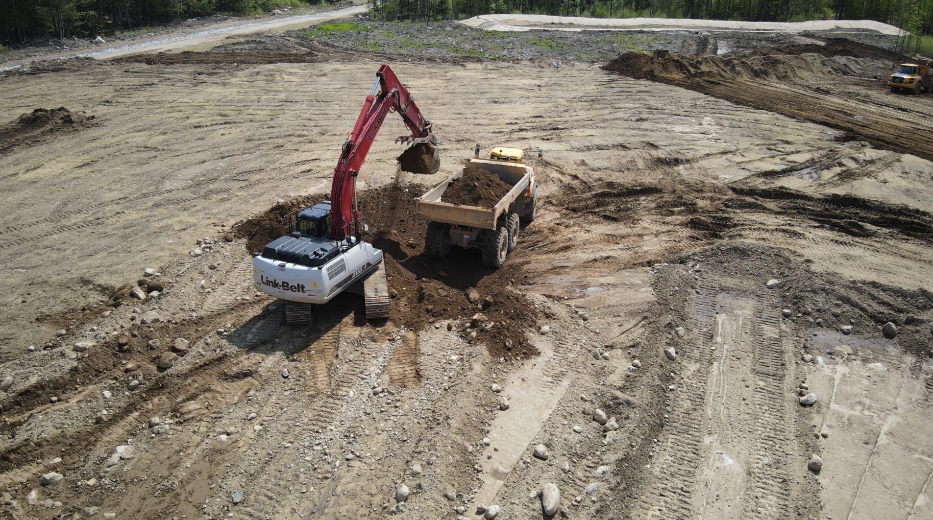 Aerial view of an excavator loading spoil into an articulated haul truck on a wide graded earthwork pad.