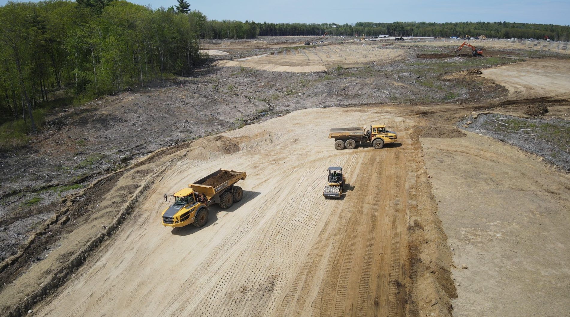 Wide aerial of an active earthwork site, two articulated haul trucks and a compactor working a graded pad.