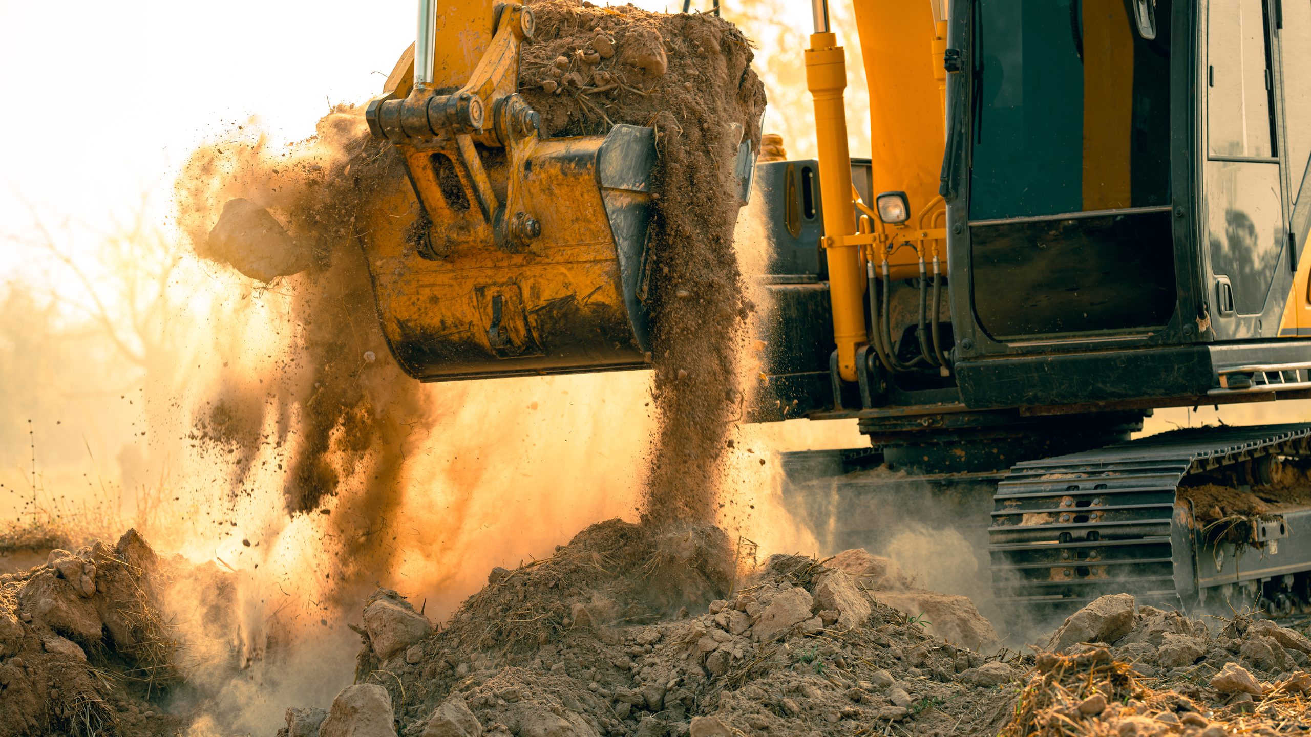 An excavator bucket dumping a load of earth in low sun, dust hanging in the air across the working face.