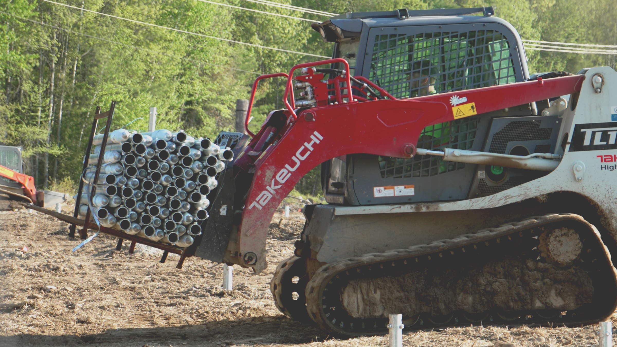 A tracked loader carrying a rack of galvanised pile sections across a cleared site, with driven piles already standing in the ground behind it.