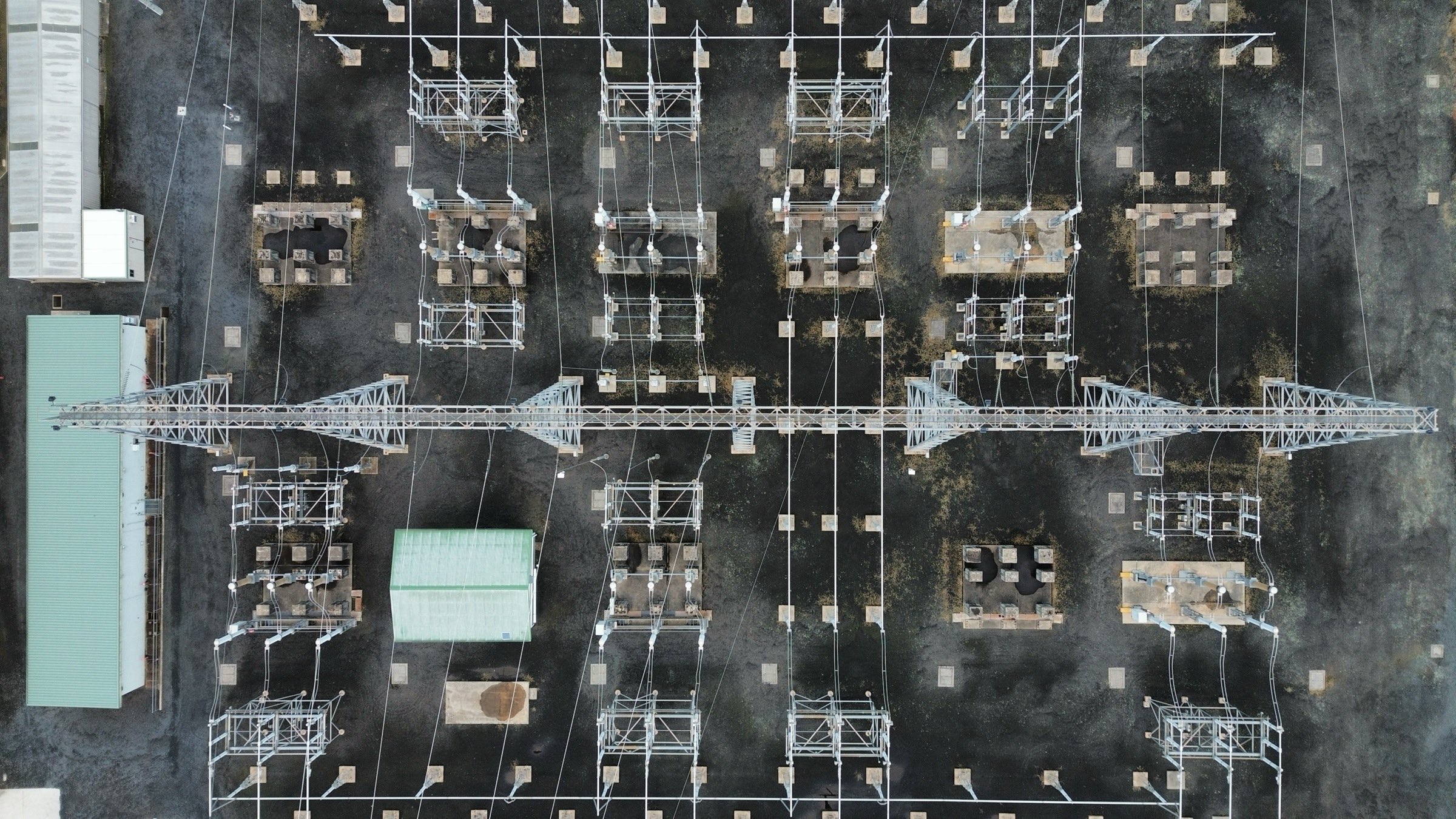 Overhead drone view of an electrical substation, transformer bays and steel gantries in a grid with conductors running across.