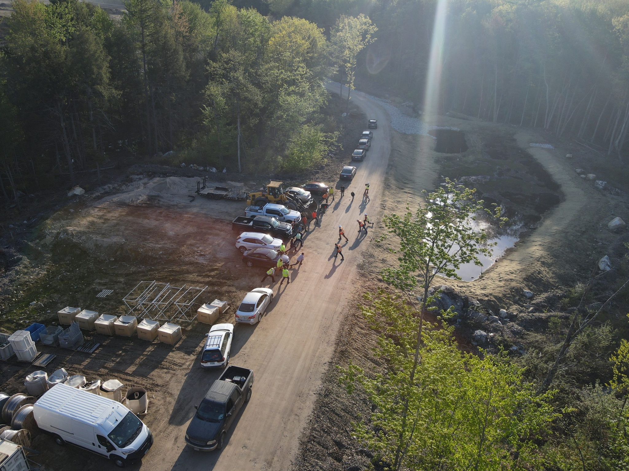 Aerial view of a site access road at shift change, a crew in hi-vis walking in past parked trucks and a laydown area.