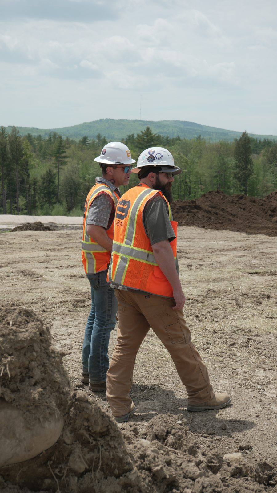 Two Brooks workers in hard hats and branded hi-vis vests walking across a graded site, wooded hills behind them.